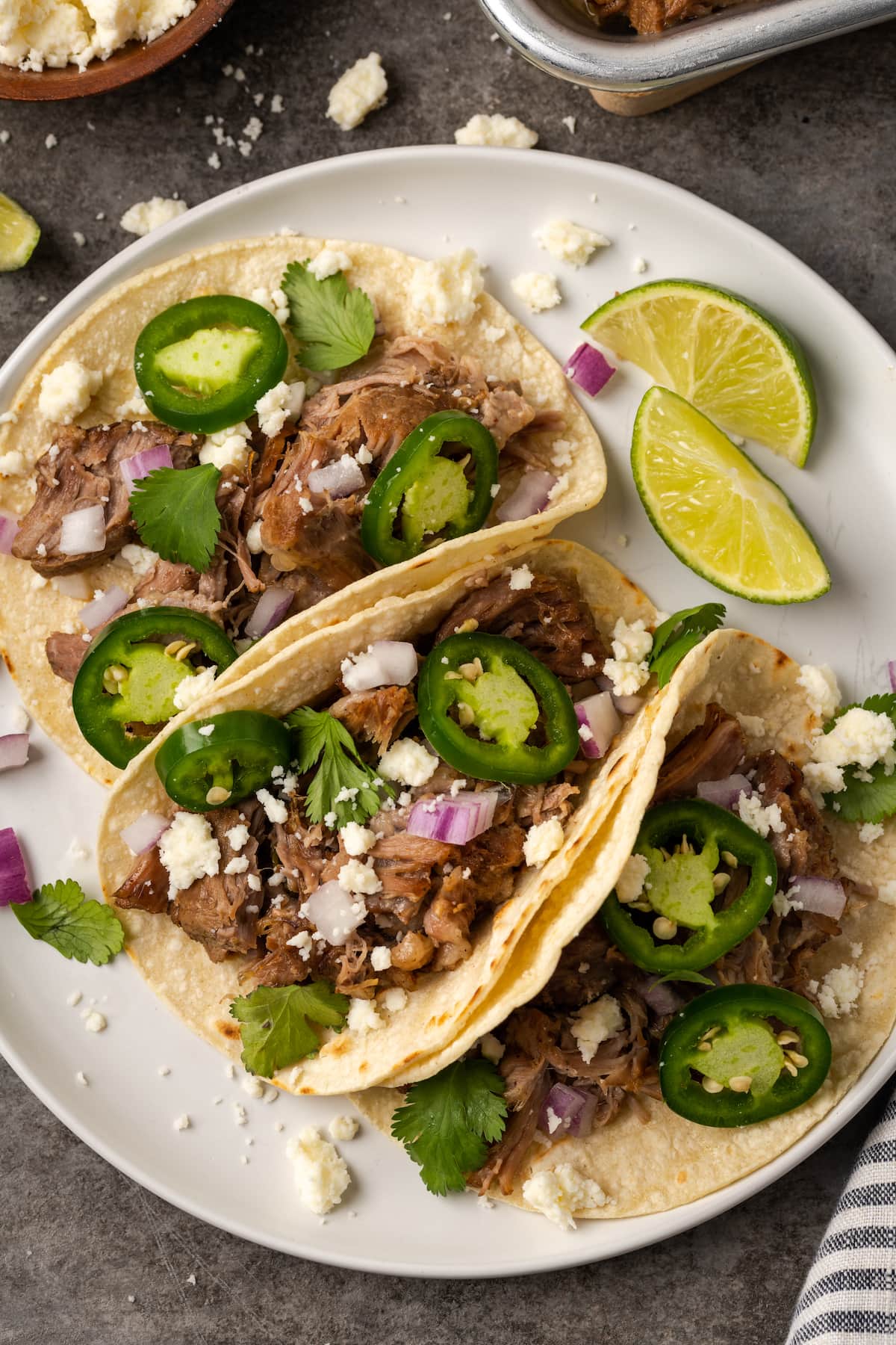 Overhead view of three pork tacos topped with jalapeños and cheese on a white plate next to lime wedges.