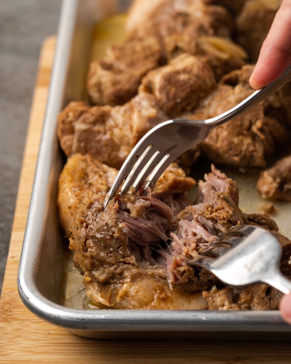 Two forks used to shred cooked pork roast on a metal baking sheet.