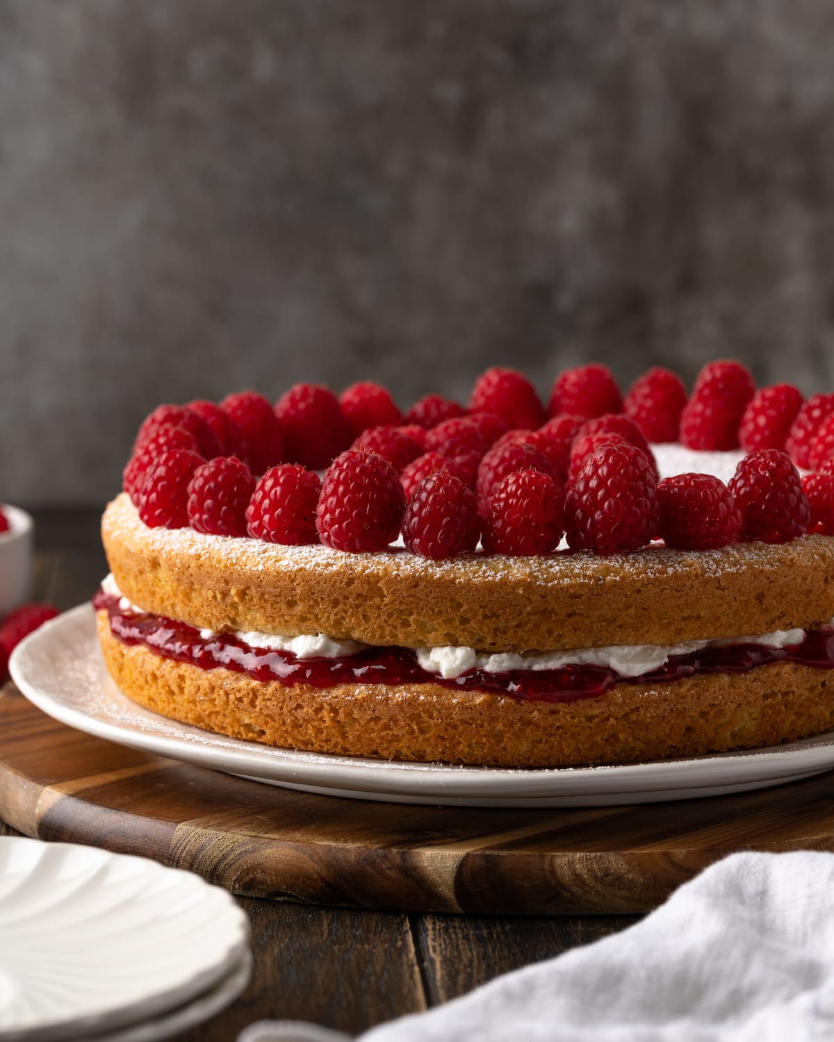 Side view of a Victoria sponge cake topped with fresh raspberries on a white plate.