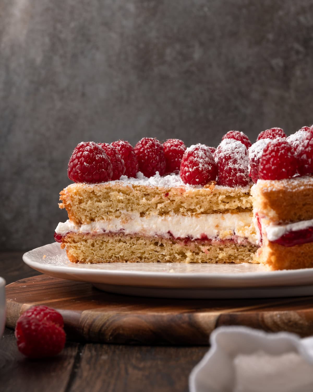 Victoria sponge cake topped with fresh raspberries on a white plate with a large slice missing.