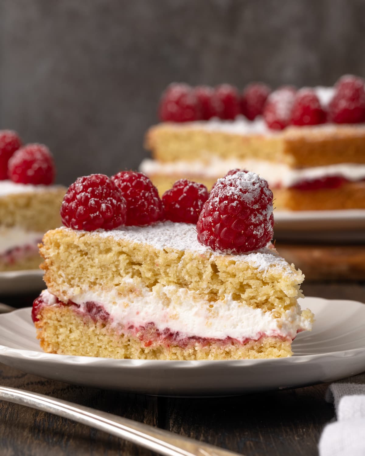A slice of Victoria sponge cake on a white plate, topped with fresh raspberries, with the rest of the cake on a plate in the background.