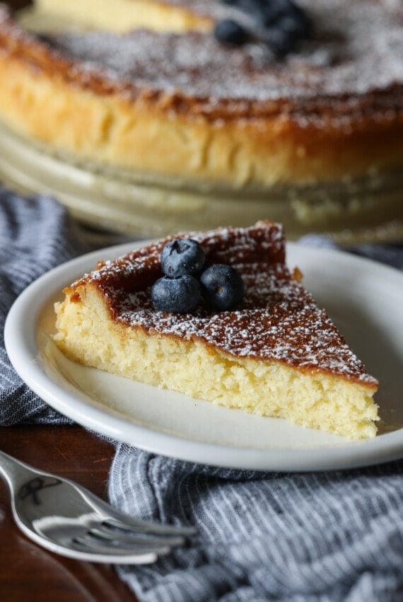 A slice of flourless white chocolate cake on a white plate dusted with powdered sugar and topped with fresh blueberries, with the rest of the cake in the background.