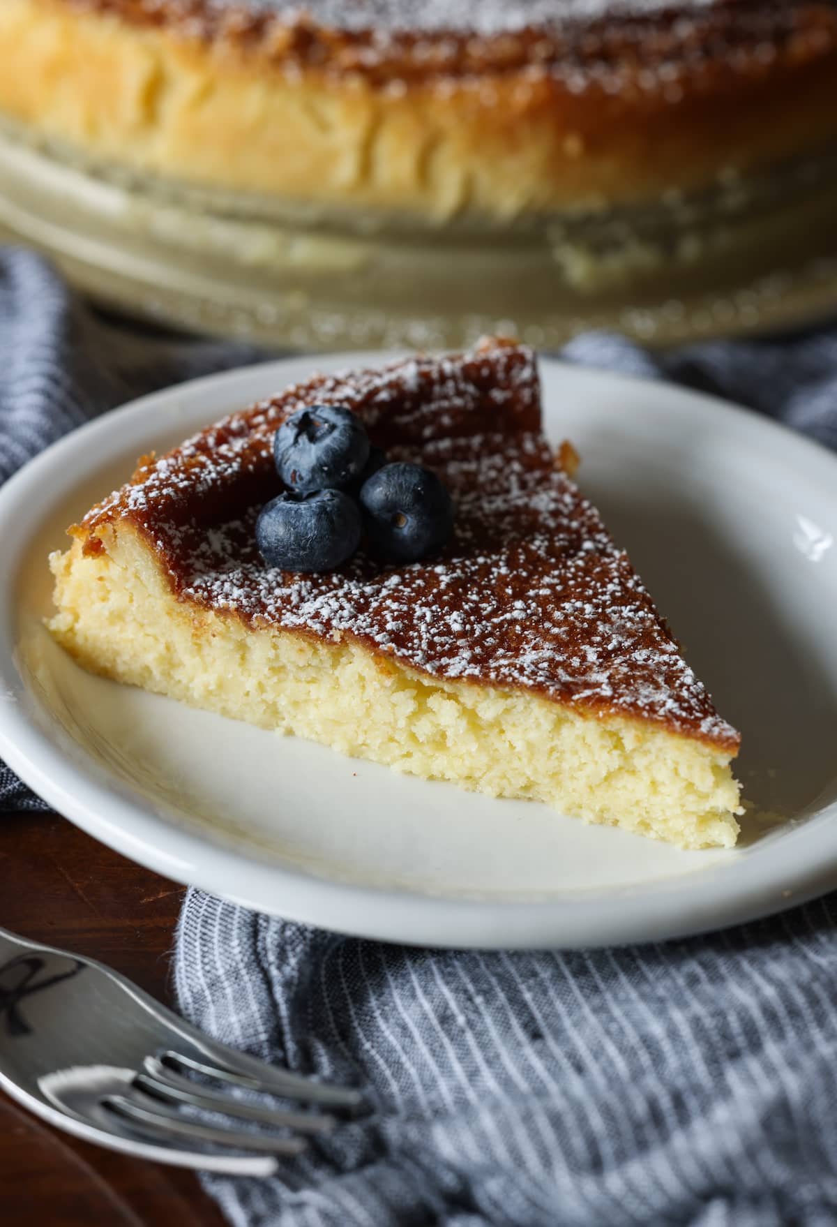 A slice of flourless white chocolate cake on a white plate dusted with powdered sugar and topped with fresh blueberries, with the rest of the cake in the background.