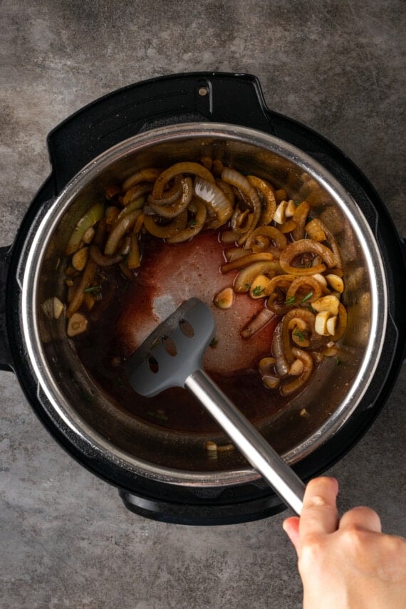 A hand uses a spatula to scrape the browned bits from the bottom of the instant pot after deglazing.