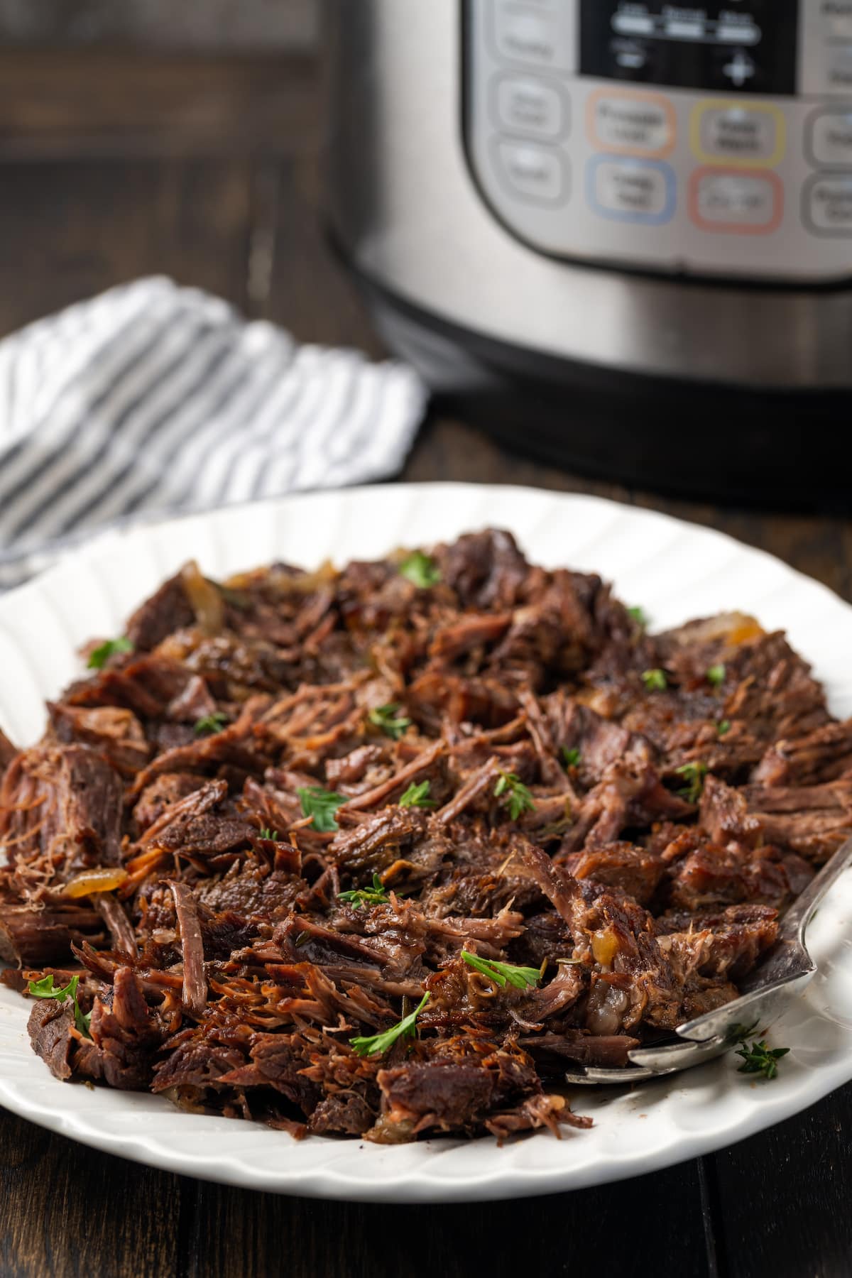 Shredded instant pot chuck roast on a white plate next to a serving spoon, with the instant pot in the background.