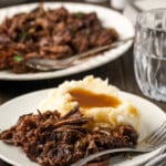 Shredded pot roast served on a white plate next to mashed potatoes covered in gravy, with more shredded roast on a plate in the background.