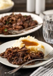 Shredded pot roast served on a white plate next to mashed potatoes covered in gravy, with more shredded roast on a plate in the background.