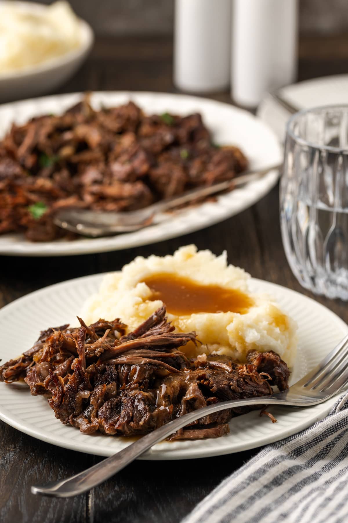 Shredded pot roast served on a white plate next to mashed potatoes covered in gravy, with more shredded roast on a plate in the background.