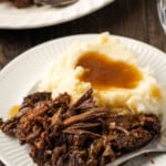 Shredded pot roast served on a white plate next to mashed potatoes covered in gravy, with more shredded roast on a plate in the background.