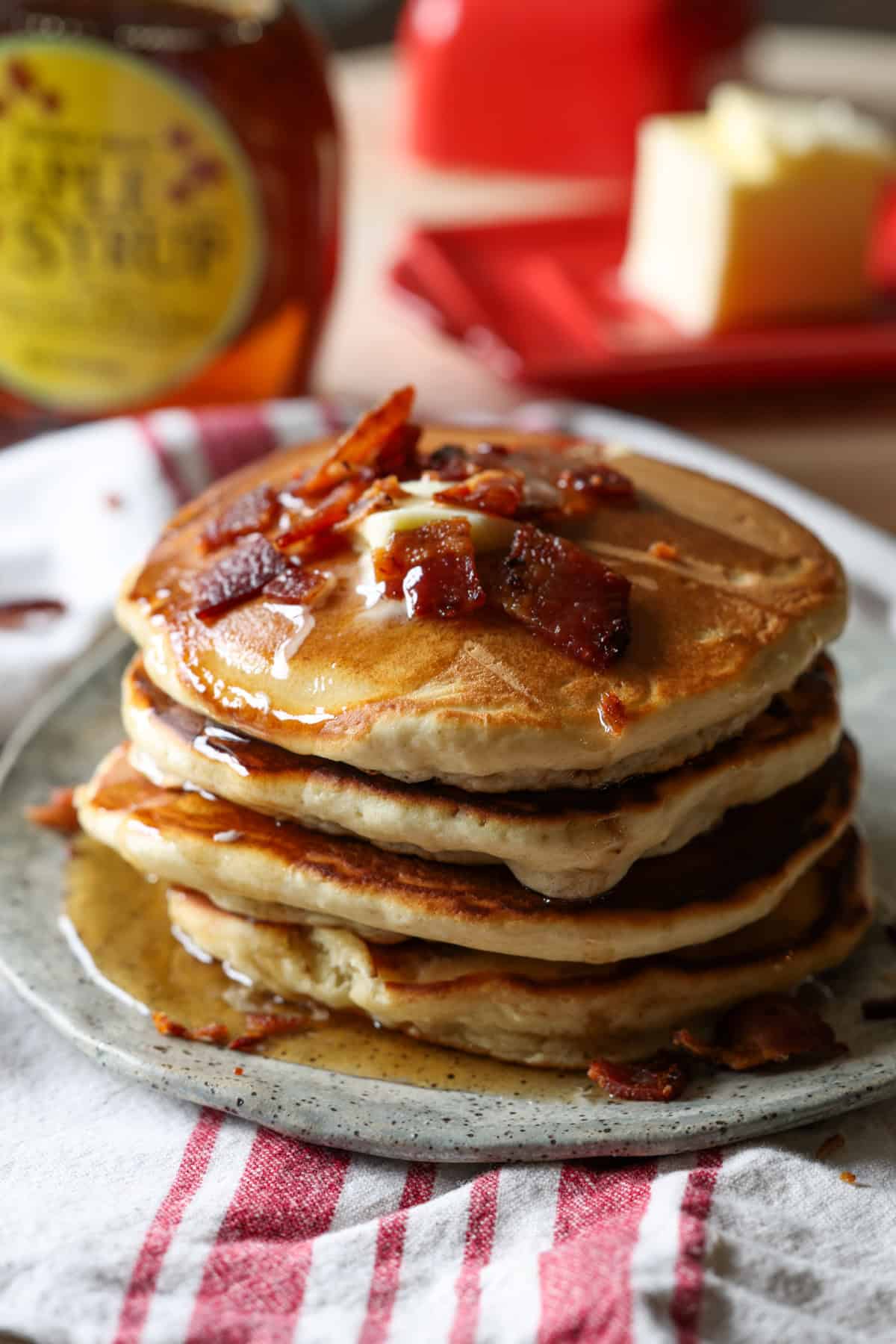A stack of pancakes with bacon and butter on top with maple syrup dripping down the sides.