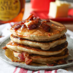 A stack of Maple Bacon Pancakes topped with butter, more chopped bacon, and maple syrup