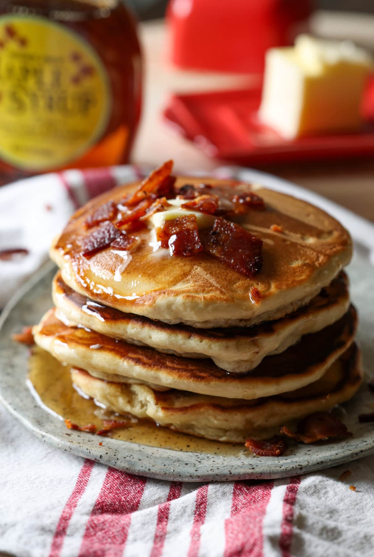 Maple Bacon Pancakes are fluffy buttermilk pancakes sweetened with brown sugar and maple syrup with crispy bacon bits inside and on top of the pancakes. A stack of pancakes with bacon and butter on top and maple syrup dripping down the sides.