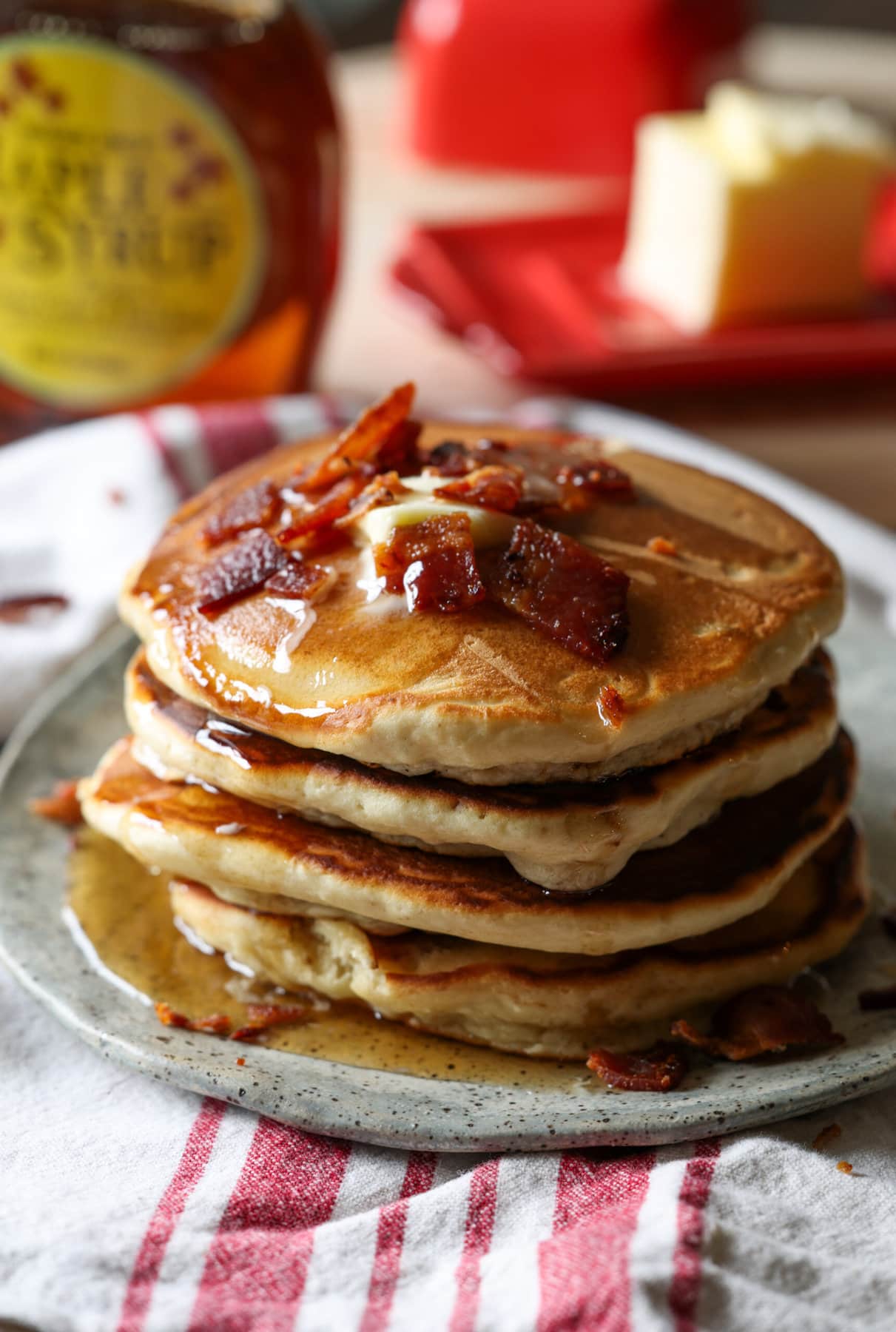 A stack of pancakes with bacon and butter on top and maple syrup dripping down the sides.