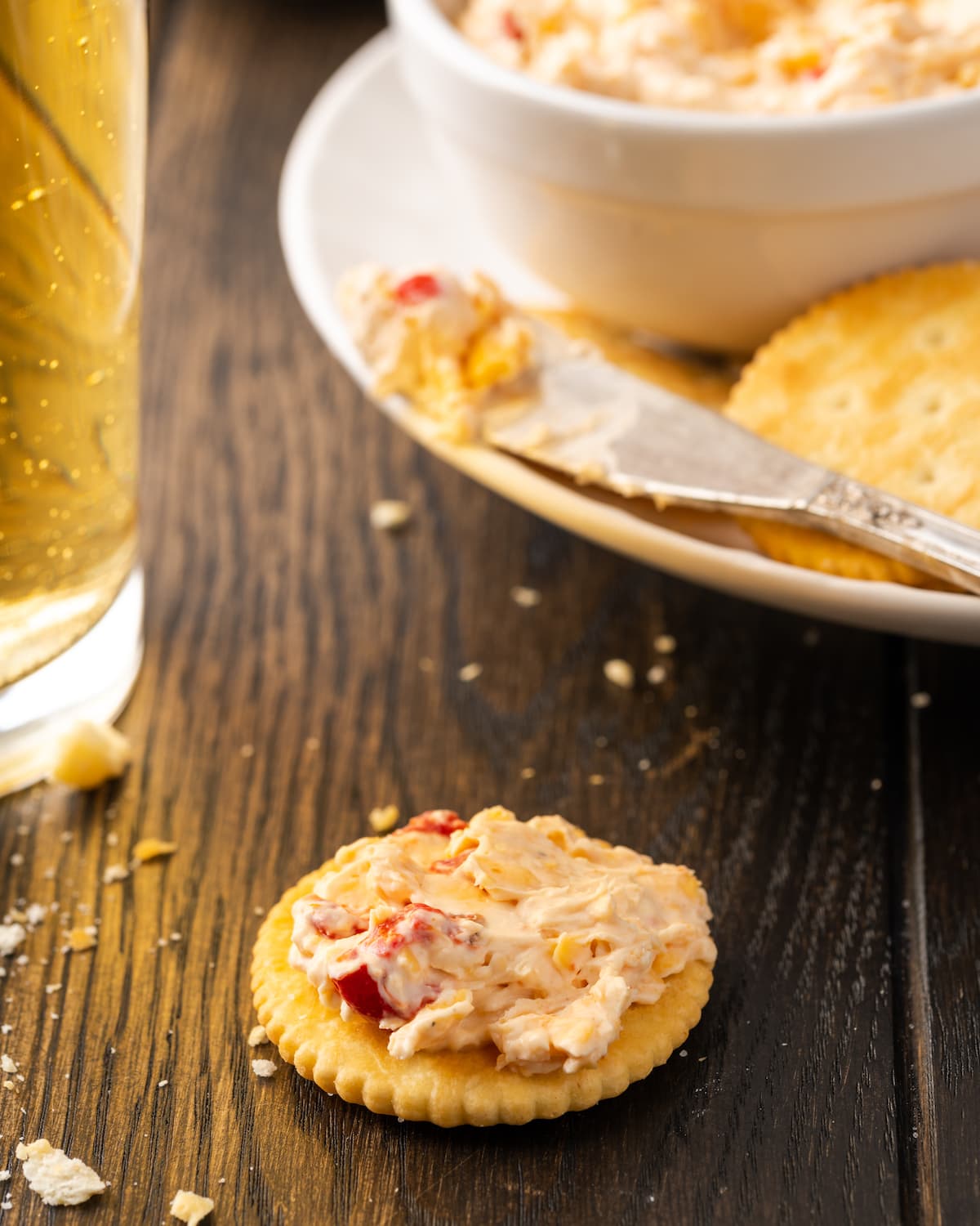 Crackers topped with pimento cheese on a tabletop with a plate of crackers and pimento cheese in the background.