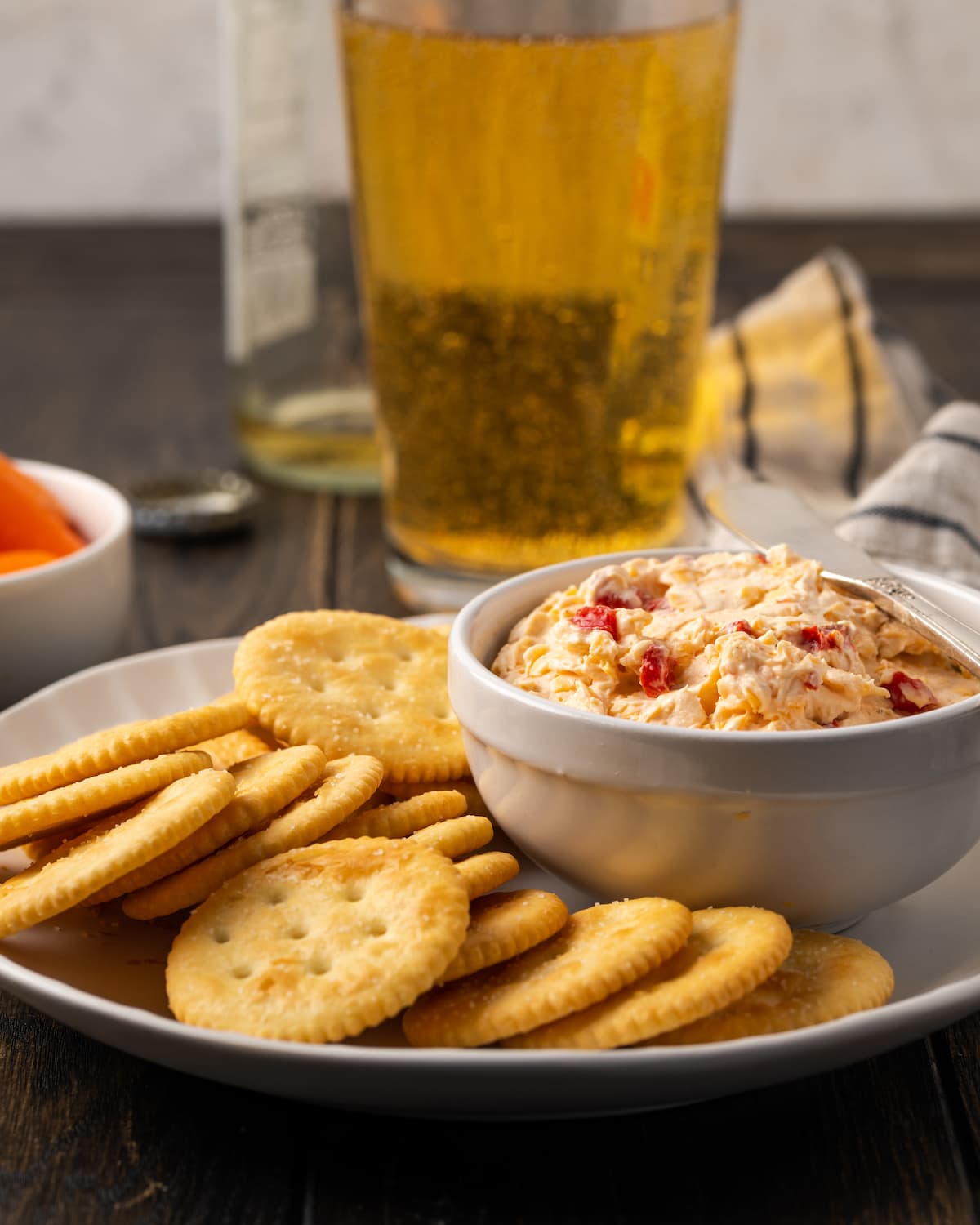 A bowl of pimento cheese is served on a plate of crackers, with glasses of beer in the background.