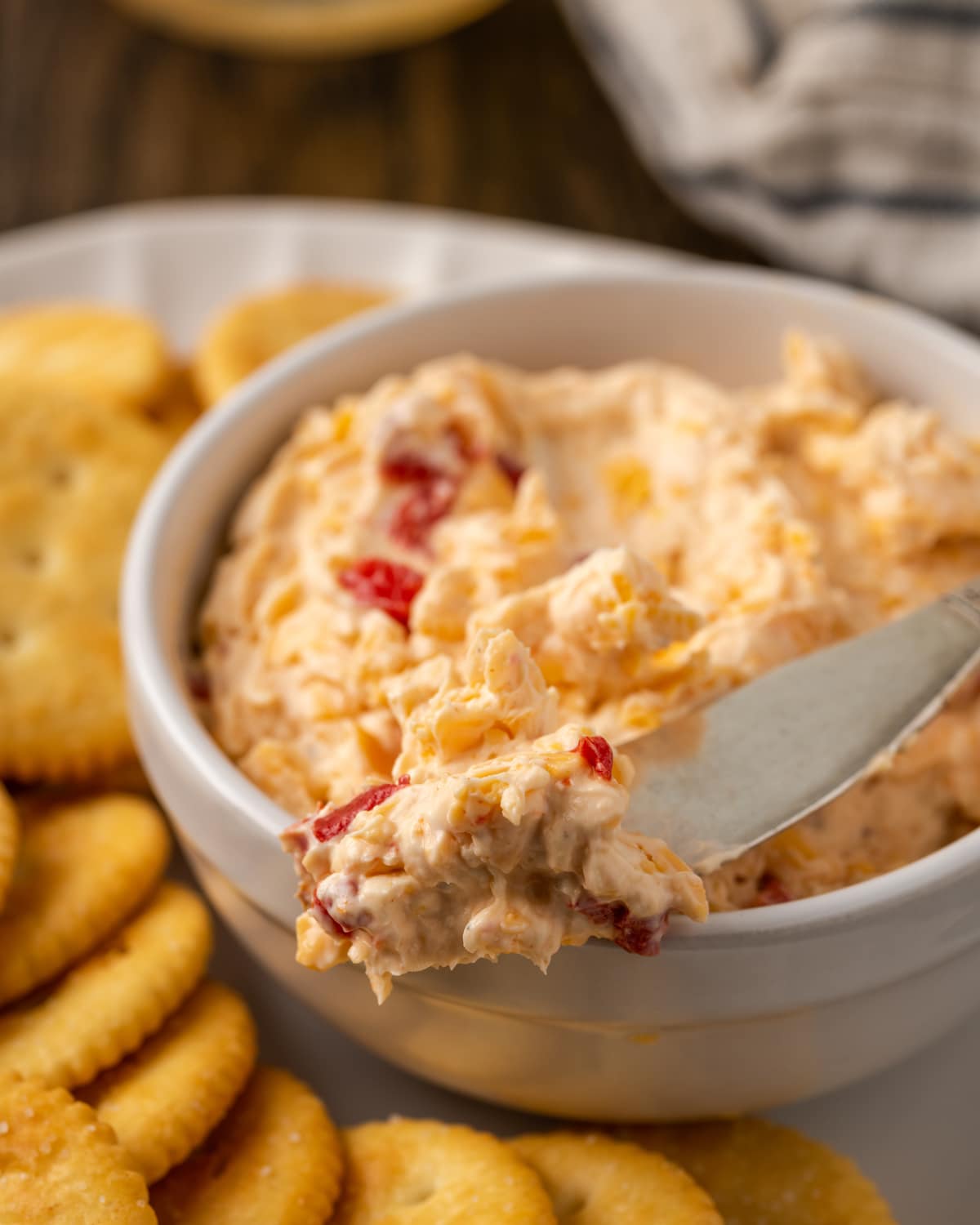 A close up of a bowl of pimento cheese with a cheese knife on a platter of crackers.