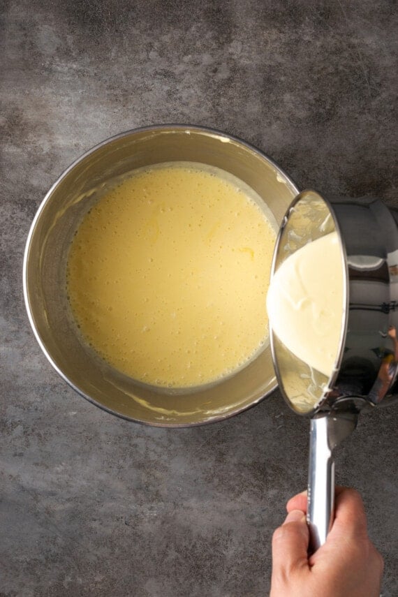 Melted white chocolate is poured from a saucepan into a metal mixing bowl with batter ingredients.