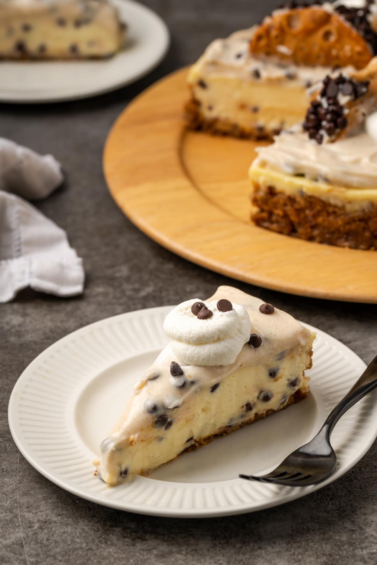 A slice of cannoli cheesecake topped with a swirl of whipped cream on a white plate next to a fork, with the rest of the cheesecake in the background.
