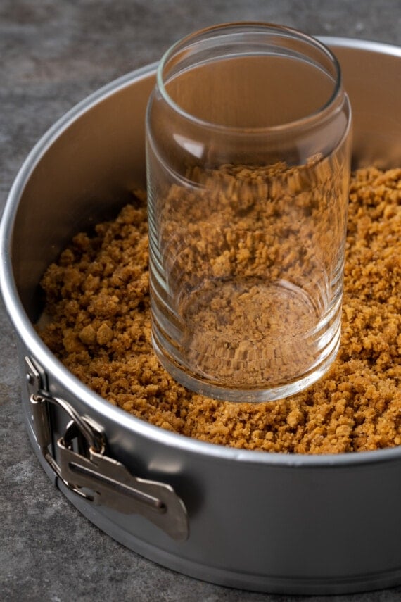 A glass jar is used to press a graham cracker crust into the bottom of a springform pan.