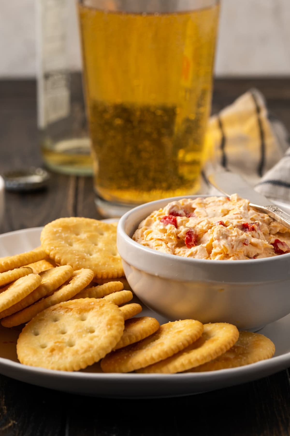 A bowl of pimento cheese served on a platter of crackers, with glasses of beer in the background.
