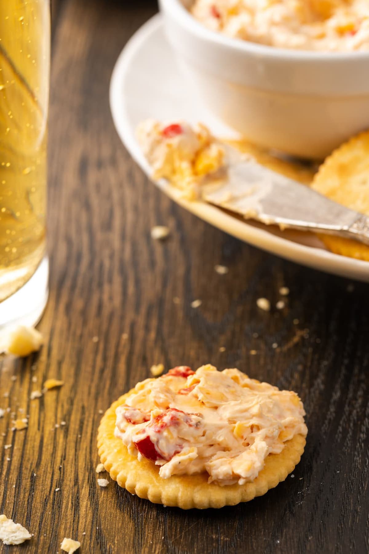 A cracker topped with pimento cheese on a tabletop with a plate of crackers and pimento cheese in the background.