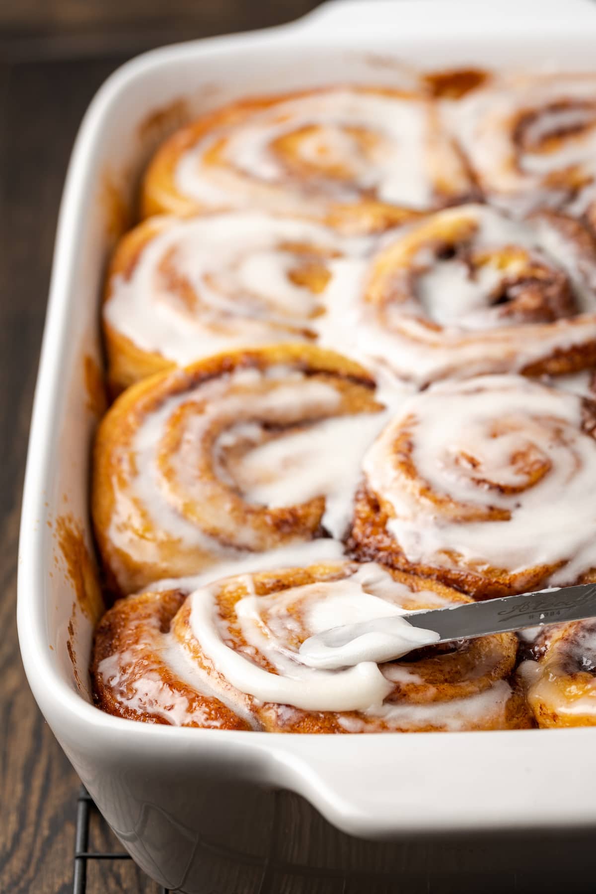 A knife spreading frosting over baked TikTok cinnamon rolls in a ceramic baking dish.