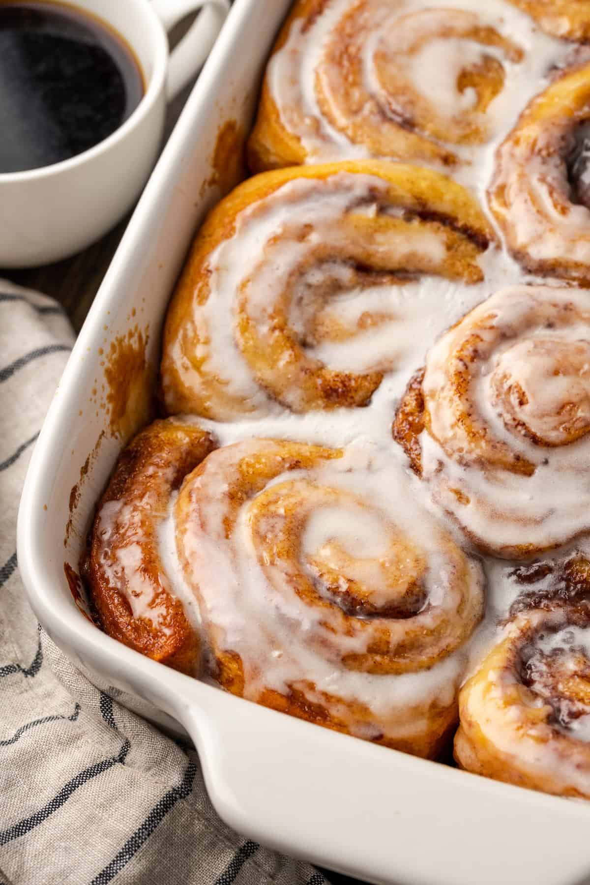 Overhead view of baked TikTok cinnamon rolls covered with icing in a ceramic baking dish, next to a cup of coffee.