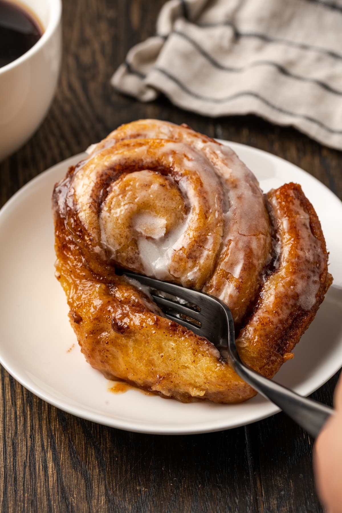 A fork cutting into the corner of a cinnamon roll on a white plate next to a cup of coffee.