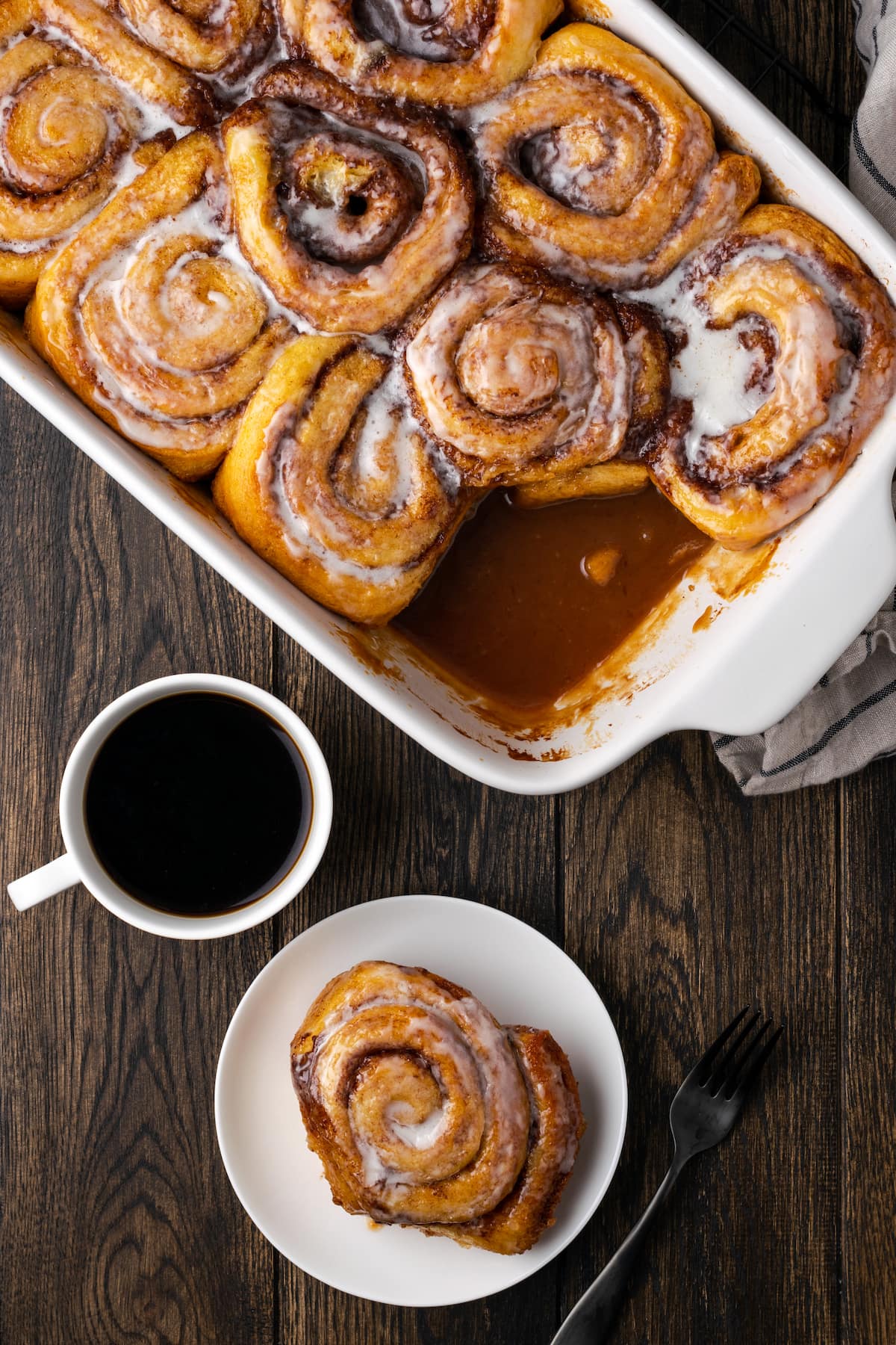 Overhead view of cinnamon rolls in a baking dish, next to one cinnamon roll served on a plate next to a cup of coffee.