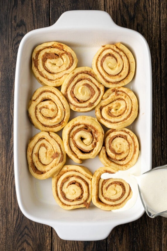 Heavy cream is poured over unbaked Grands cinnamon rolls in a white ceramic baking dish.