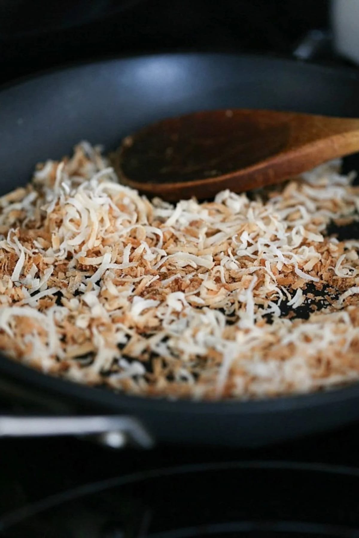 toasted coconut in a black skillet with a wooden spoon