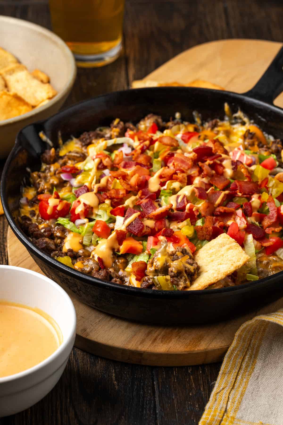 Cheeseburger dip in a skillet on a countertop next to bowls of burger sauce and chips for dipping.