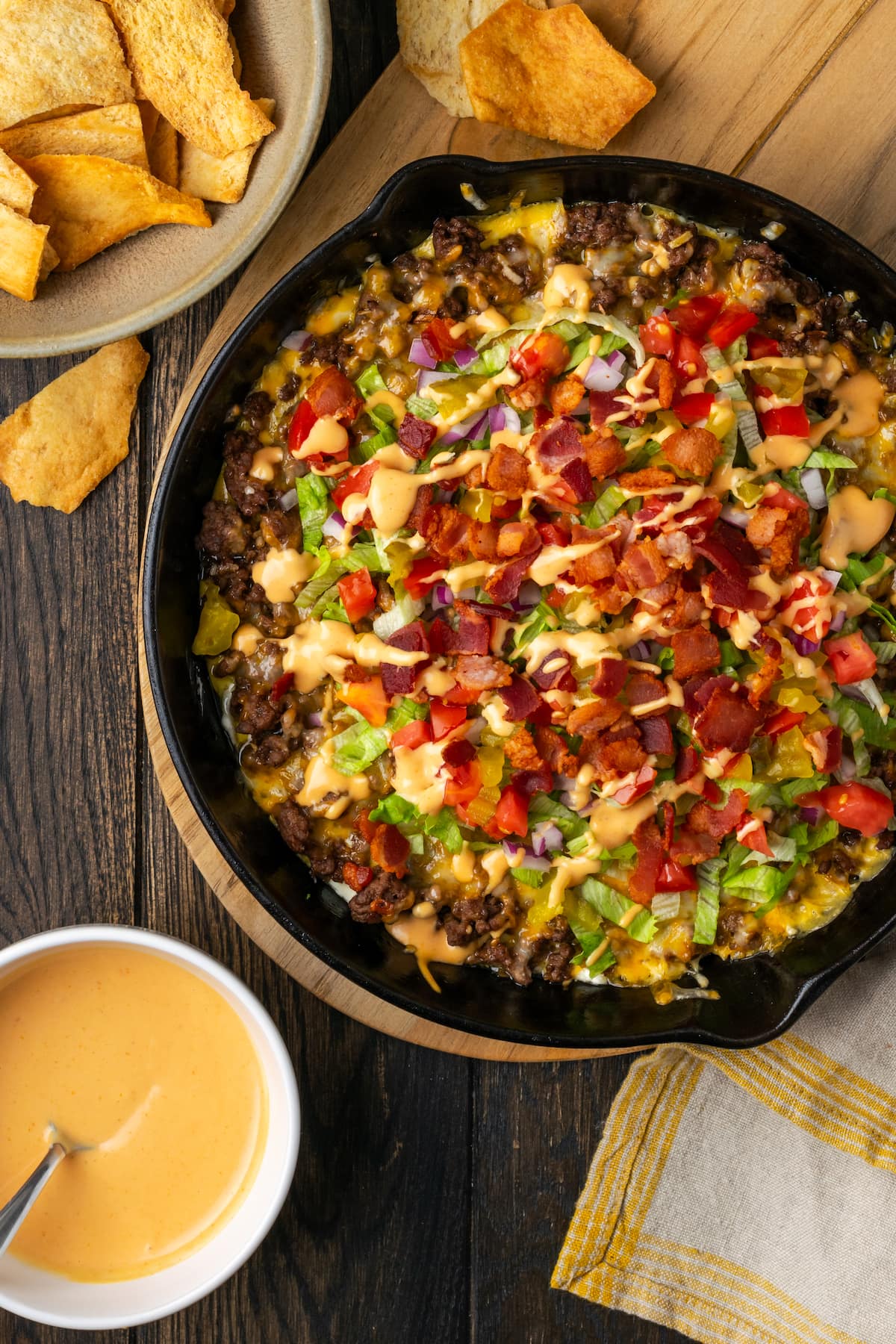 Overhead view of cheeseburger dip in a skillet on a countertop next to bowls of burger sauce and chips for dipping.