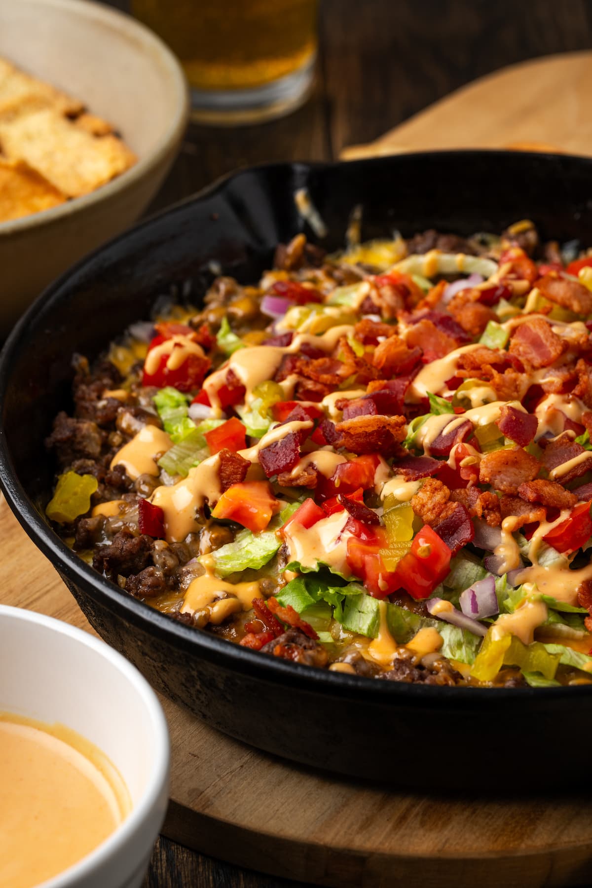 Cheeseburger dip in a skillet on a countertop next to bowls of burger sauce and chips for dipping.