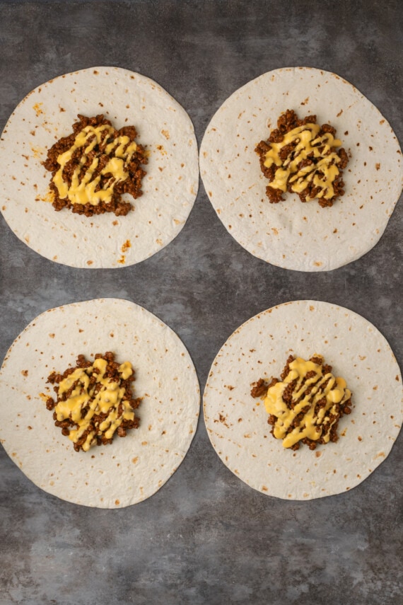 Overhead view of four partially assembled crunch wraps topped with meat and nacho cheese.