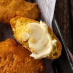 Close up of hot water cornbread in a basket, with one fritter broken in half and spread with butter.