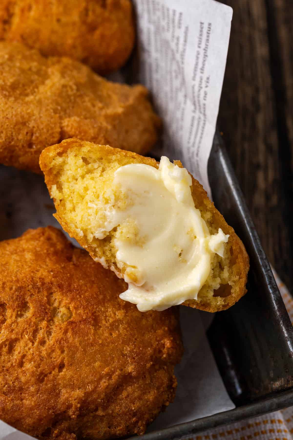 Close up of hot water cornbread in a basket, with one fritter broken in half and spread with butter.