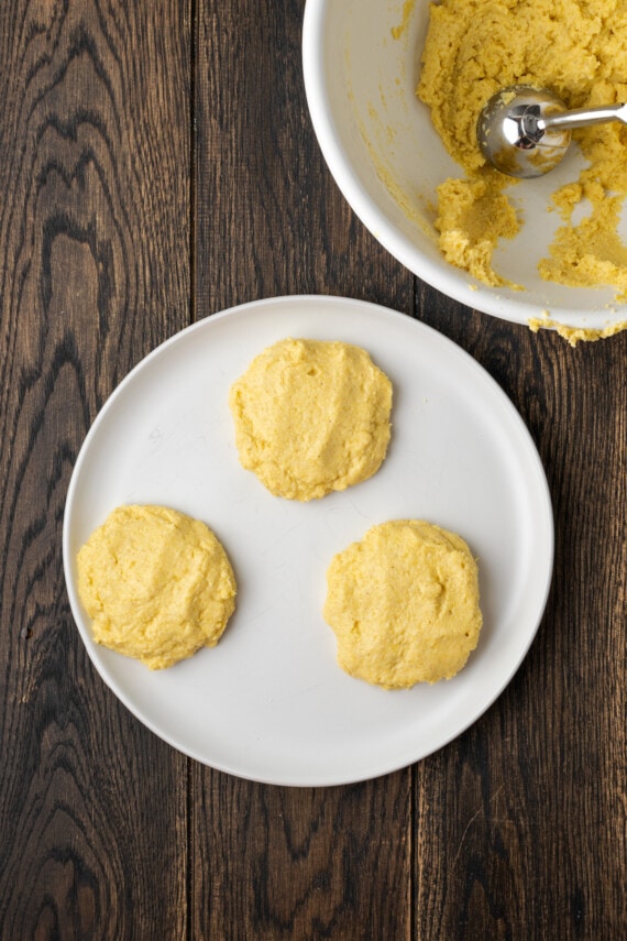 Three portions of cornbread dough on a plate next to a mixing bowl.