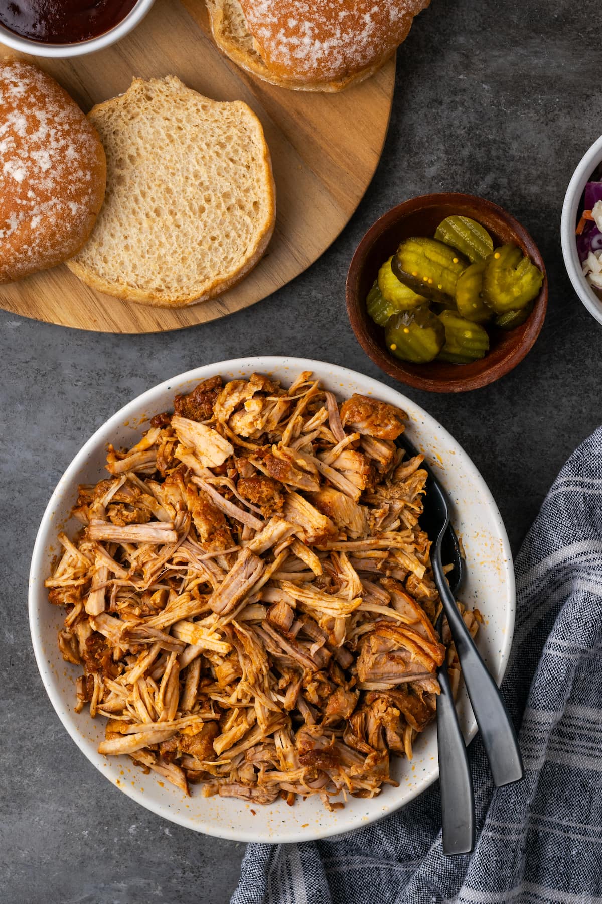 Overhead view of a bowl of Instant Pot pulled pork next to buns cut in half on a wooden cutting board, and a small bowl of pickles.