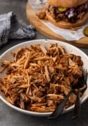 A bowl of Instant Pot pulled pork with two black forks, with a pulled pork sandwich in the background.