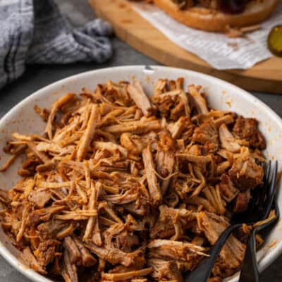 A bowl of Instant Pot pulled pork with two black forks, with a pulled pork sandwich in the background.