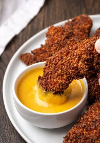 A hand dipping a pecan crusted chicken tender into a small bowl of dipping sauce, next to more chicken tenders on a plate.