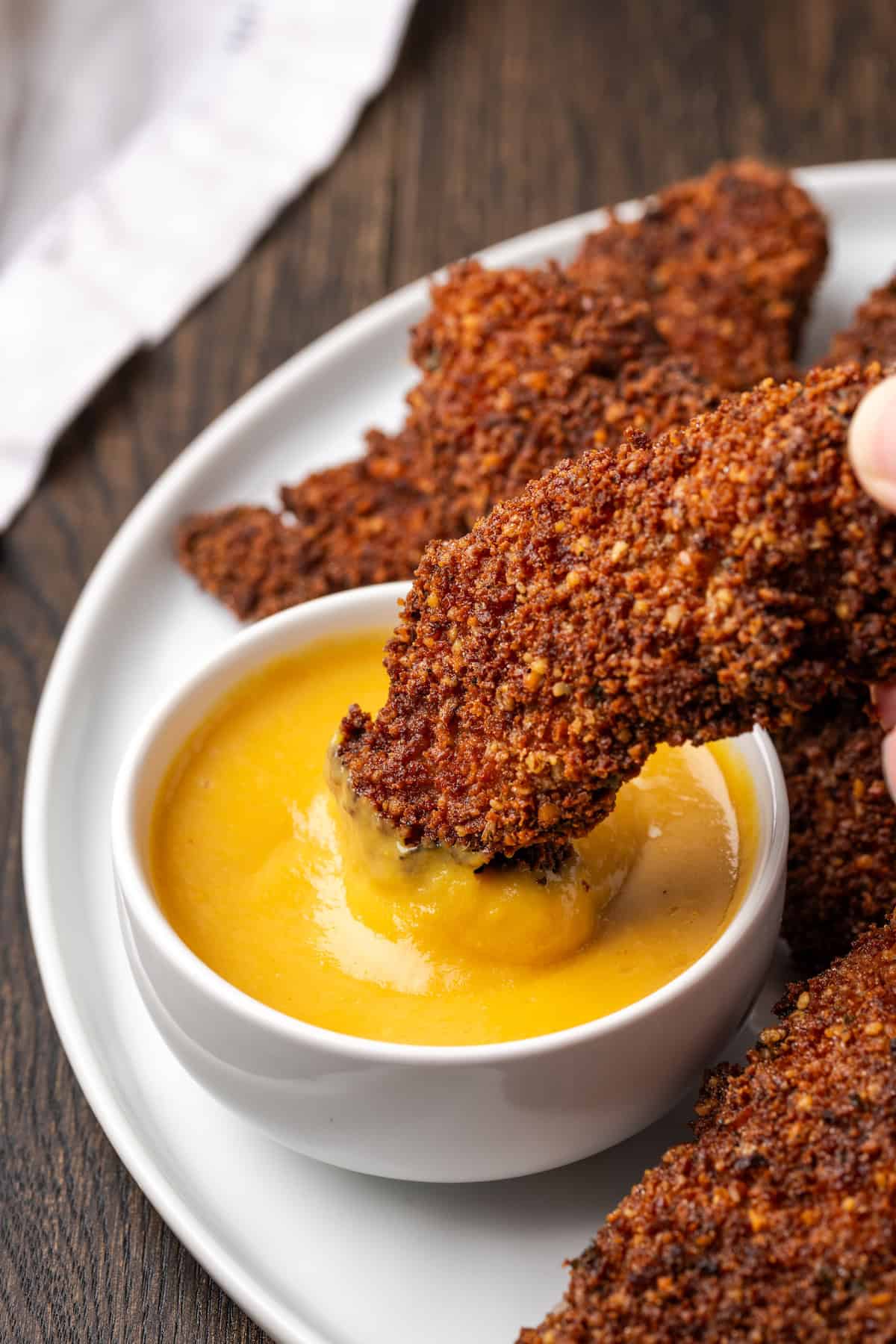 A hand dipping a pecan crusted chicken tender into a small bowl of dipping sauce, next to more chicken tenders on a plate.