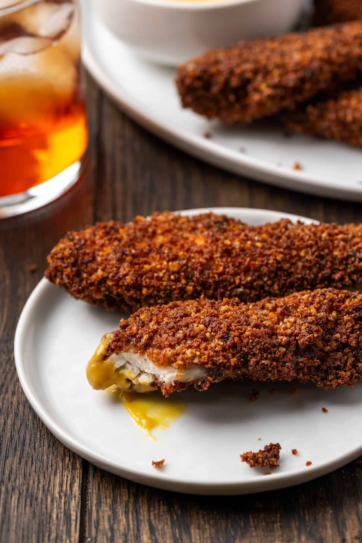 Two pecan crusted chicken tenders on a white plate, with a bite missing from one chicken tender.