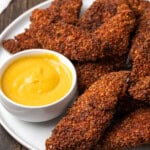 Pecan crusted chicken tenders on a plate next to a bowl of dipping sauce.