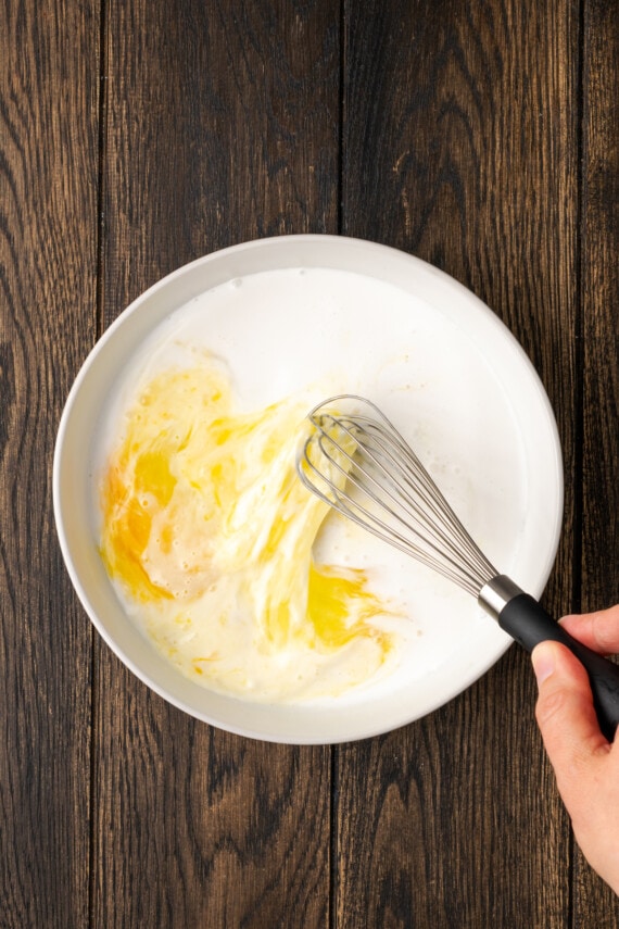 A hand uses a whisk to combine buttermilk and eggs in a bowl.