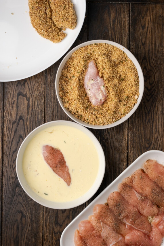 Overhead view of a breading station, with chicken tenders being dipped into bowls of buttermilk and pecan breading.