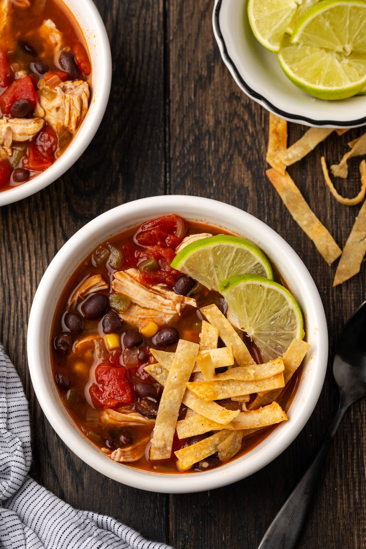 Overhead view of a bowl of chicken tortilla soup garnished with lime wedges and crispy tortilla strips, next to bowls of more soup and lime wedges.