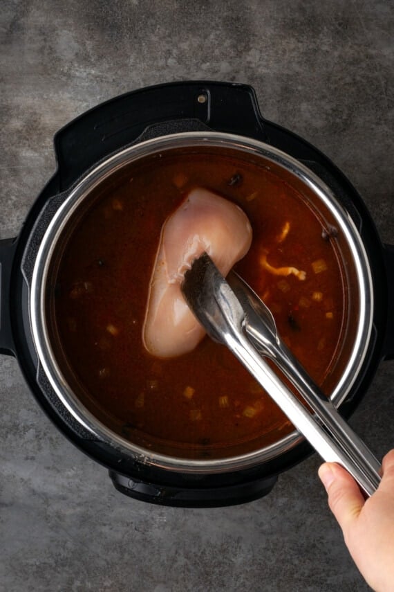 Tongs are used to lower a raw chicken breast into tortilla soup in the Instant Pot.