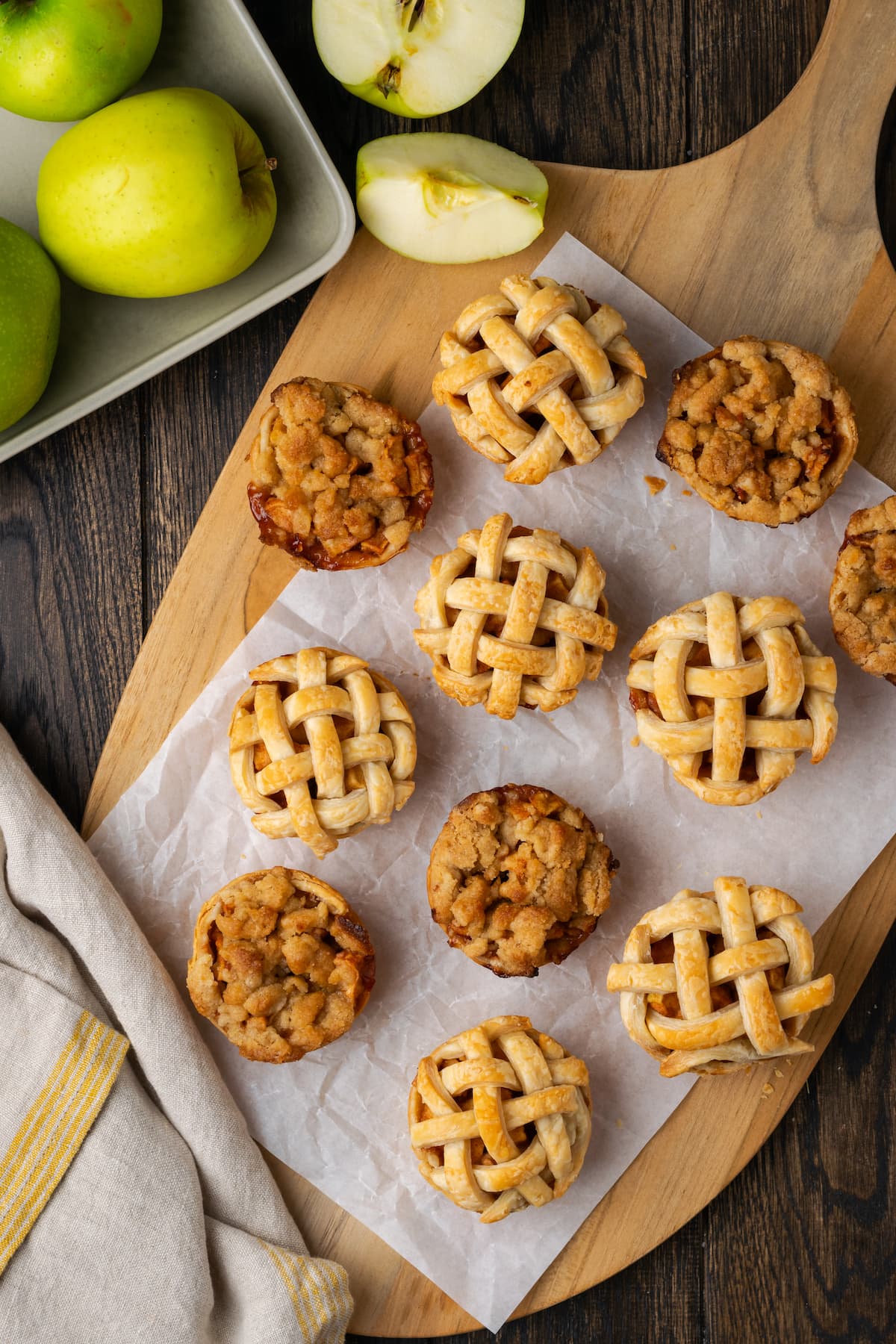 Overhead view of mini apple pies on a wooden board lined with a piece of parchment paper, next to granny smith apples in a bowl.
